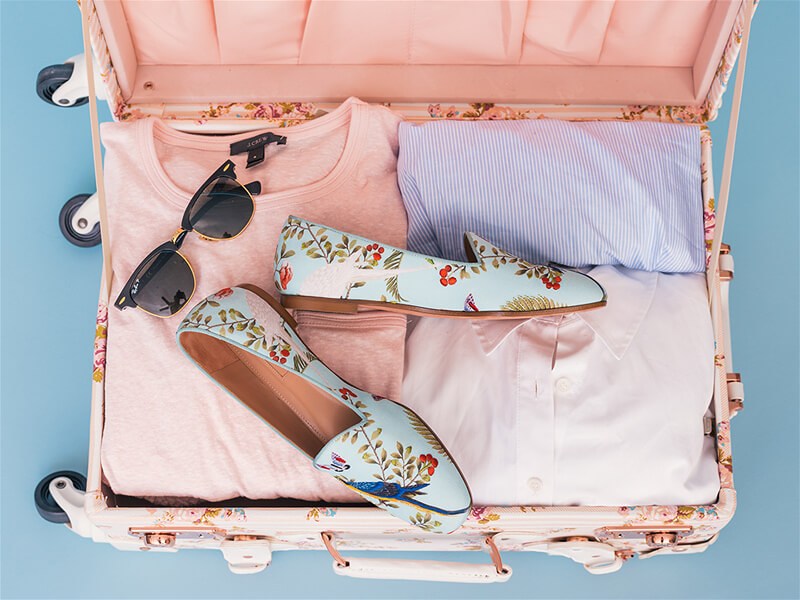 mala cor de rosa, com algumas peças de roupa de cor clara dentro da mala, um óculos de sol sobreposto em cima das roupas junto com um par de sapatos floridos