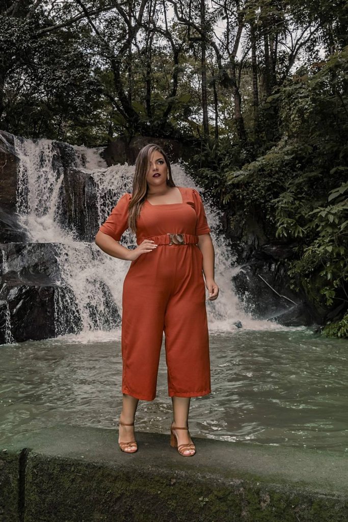 Mulher usando conjunto de calça e blusa laranjada, ao fundo cachoeira
