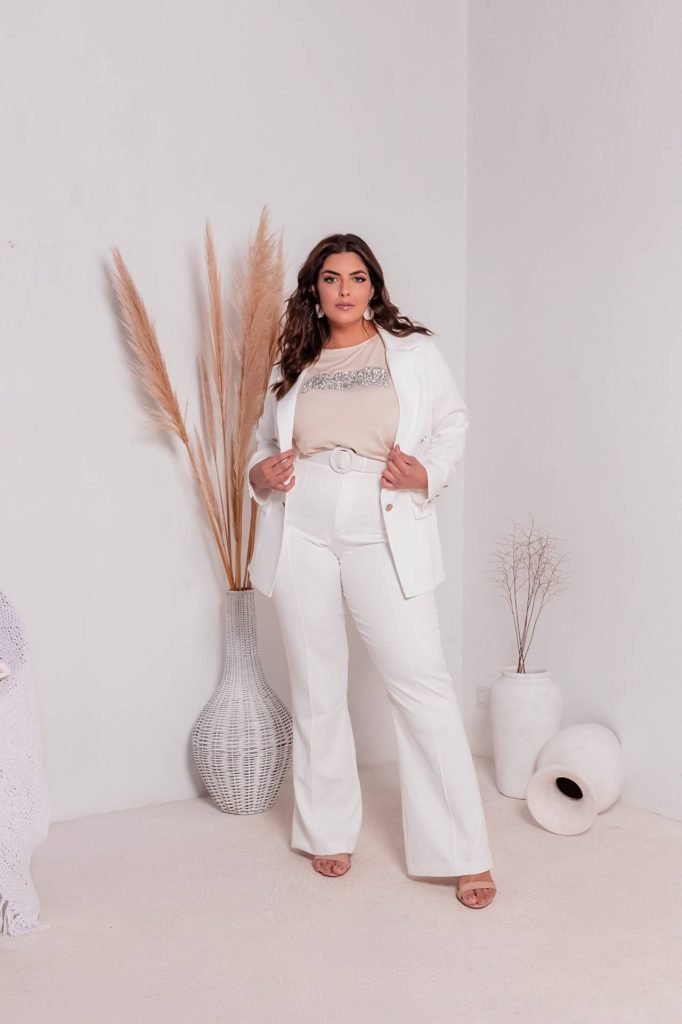 Modelo mulher usando conjunto de calça e blazer na cor branco, blusa no tom bege por baixo. Vasos decorativos no chão